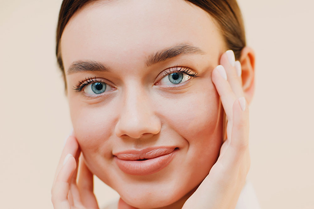 A woman with blue eyes and a gentle smile touches her face with both hands against a soft beige background, conveying a sense of natural beauty and calm.